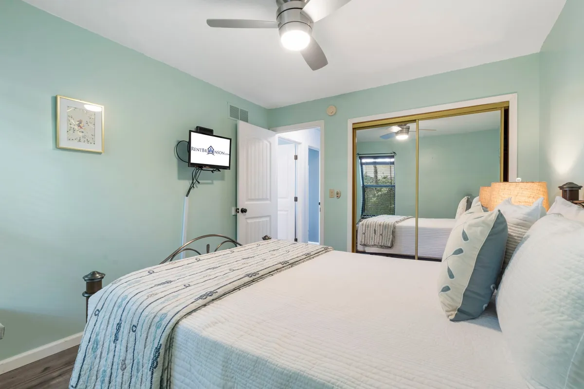 A tranquil bedroom furnished with a bed adorned with decorative pillows, a wall-mounted TV, and mirrored closet doors. The space is calm, inviting, and suited to winding down after a day of exploration.