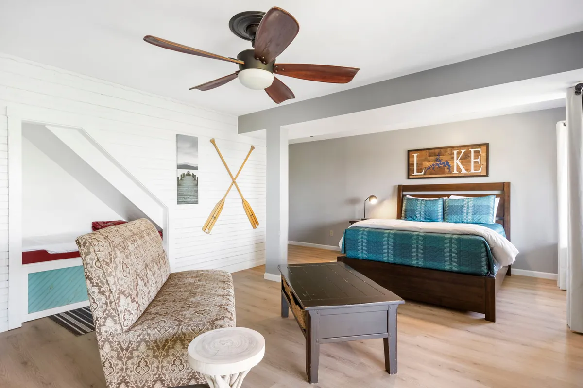 A welcoming bedroom featuring a bed with teal bedding, wooden wall art reading 'LAKE', ceiling fan, decorative oars, and a sitting area with a patterned chair. The space is bright with natural light and styled with cozy, lakeside decor.