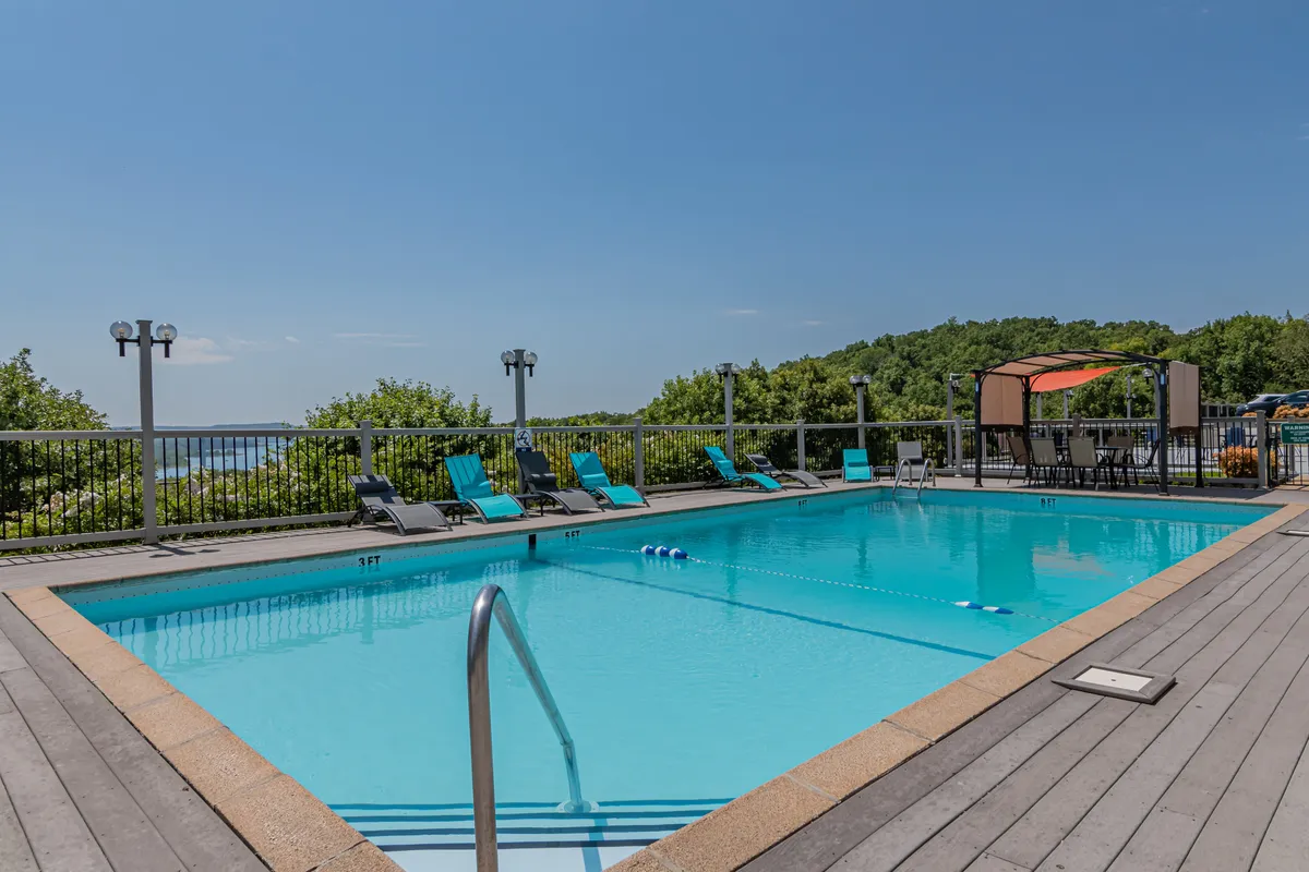 An inviting exterior view of a resort-style swimming pool with lounge chairs and a canopy, surrounded by lush greenery and a clear blue sky. The pool area looks ready for summer fun and family relaxation.