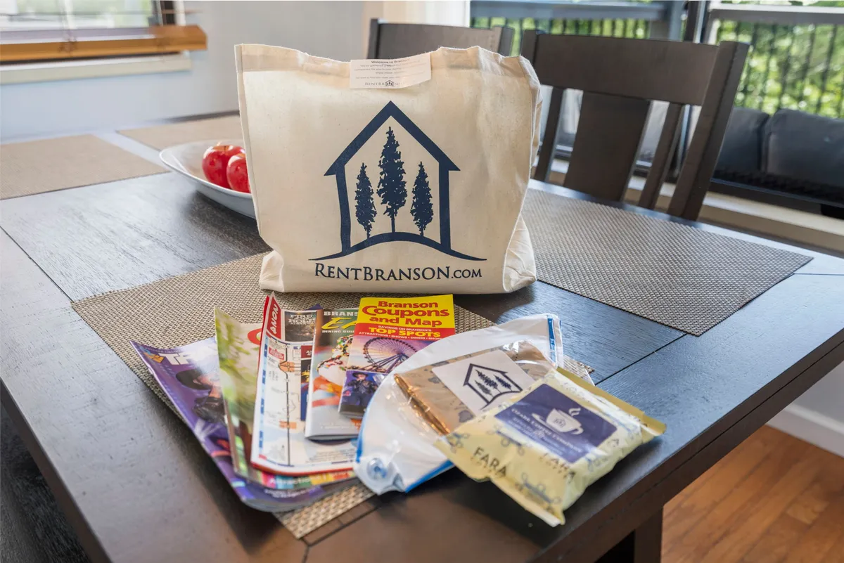 A wooden dining table with a bowl of red apples sits near a stack of brochures and a canvas bag with a local logo, evoking a sense of planning and sharing meals. The scene feels inviting and hints at both mealtime and the excitement of exploring local attractions.