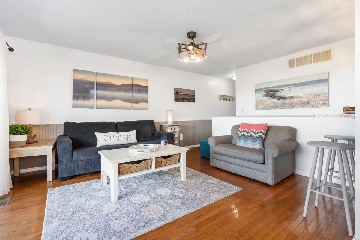A cozy living room scene with a sofa, coffee table, artwork on the wall, and a floor lamp. Morning sunlight brightens the space, making it perfect for relaxing or gathering with family.