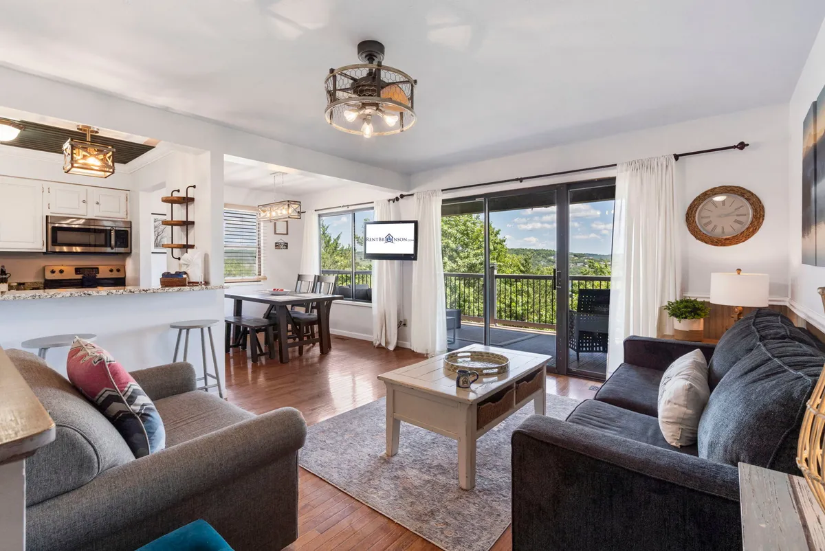 The image showcases a spacious, open-concept living and dining area featuring a cozy sofa and a dining table, all bathed in natural light from large windows that offer stunning views of Table Rock Lake. The decor is modern yet warm, with a prominent decorative clock adding style, while the windows frame the tranquil Ozarks landscape, inviting guests to relax and enjoy the scenery from indoors.
